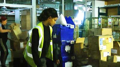 distance photo of team 3 with a young uniformed woman in focus surrounded by boxes of stock bending over and packaging one box ready for transport to the basement