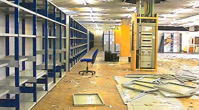 distant internal corner photo of large retail shop premises consisting of racking and shelving on the left and the shop floor litered with partially dismantled racking and shelves to the right and a lonesome blue office chair offset to the left of a large central white building column support