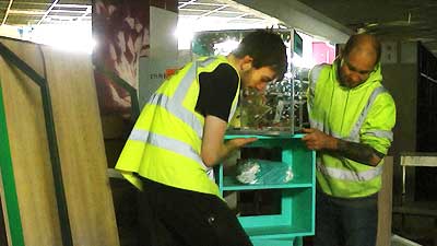 close up of hi vis uniformed team 4 carrying a small bright cyan blue retail display cabinet with a young man and an older man at the sides