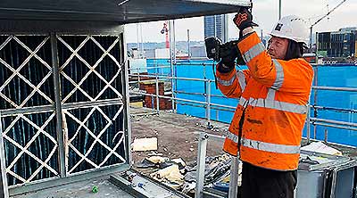 Close up photo of a howarth site expert dressed in a white hard hat and orange high visibility jacket and black trousers standing center picture using a cordless screwdriver facing upwards to dismantle a very large air conditioning ducting venting system